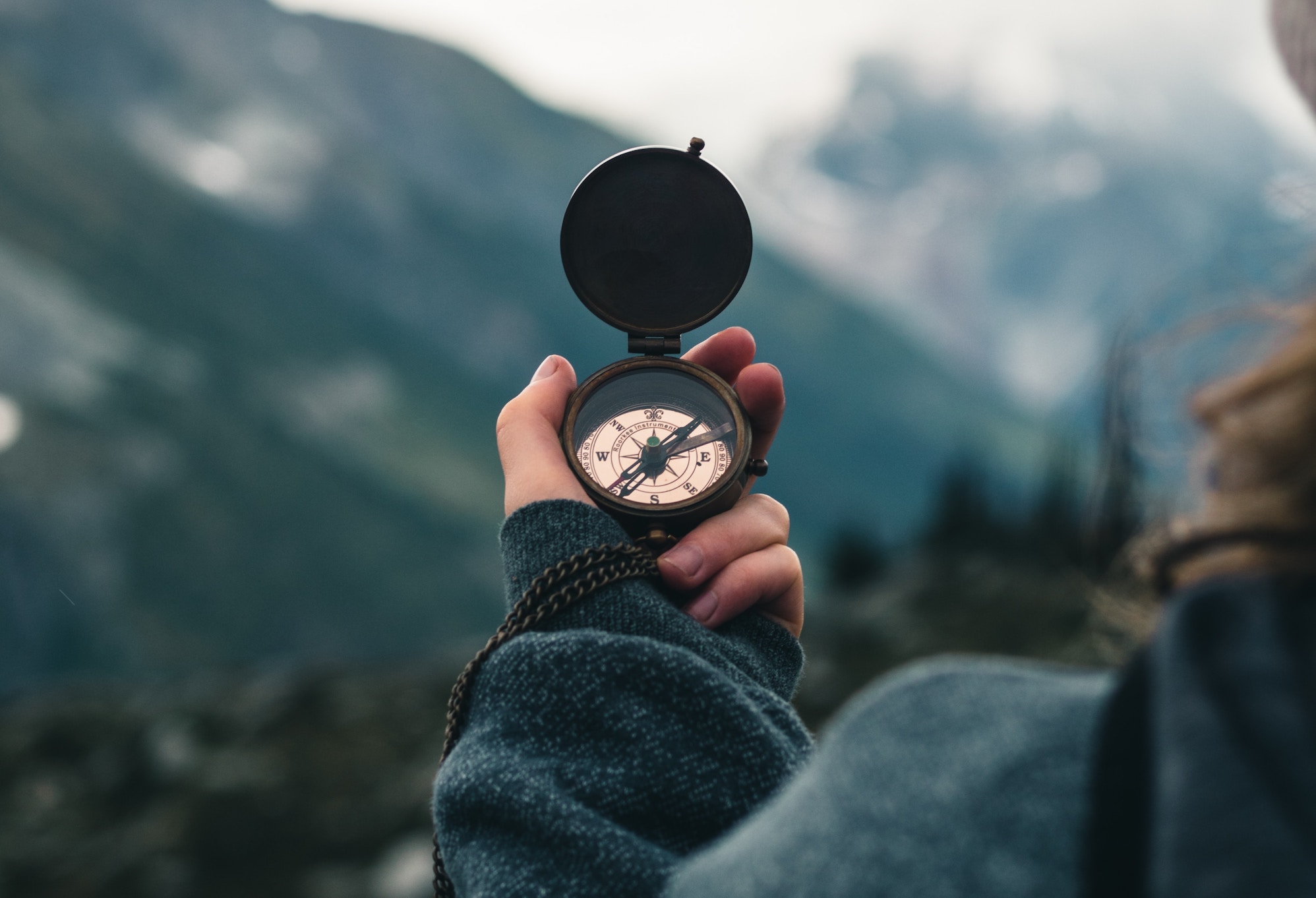 Supporting image: Person in a blue jumper holds a compass up in front of blurred mountains.